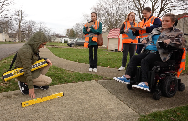 A member of the accessibility audit team uses a level to determine the slope of a sidewalk while other members watch, one taking notes on a clipboard.