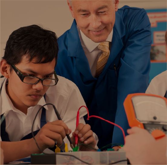A teacher watching a young boy with glasses and a young girl working together on a electronic device in a class.