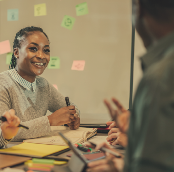 Young adult woman sitting at a desk discussing things with colleagues. Post-it notes are around them to show brainstorming took place.