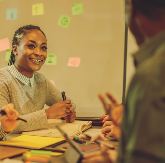Young adult woman sitting at a desk discussing things with colleagues. Post-it notes are around them to show brainstorming took place.