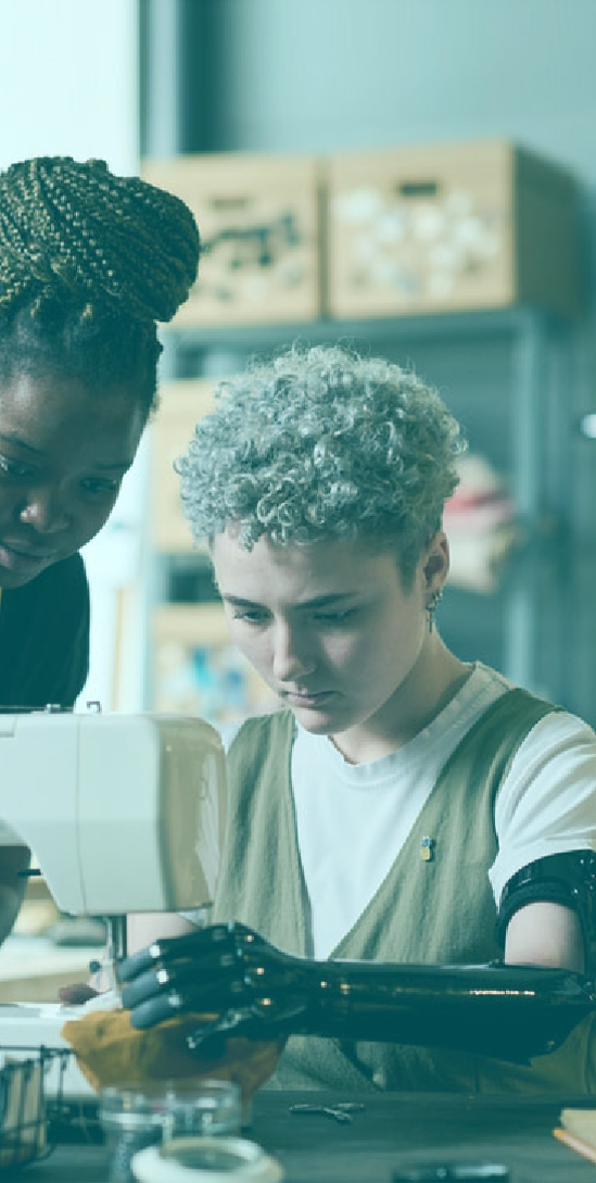 Young adult with prosthetic arm using a sewing machine with a mentor standing over and watching