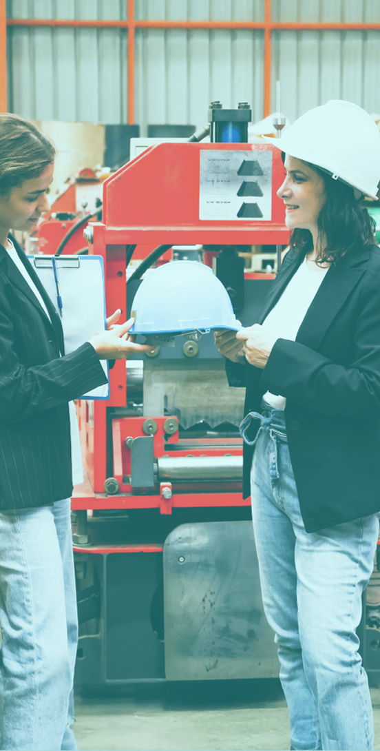 A woman handing a hard hat to a younger woman in front of 2 large machines in a work room